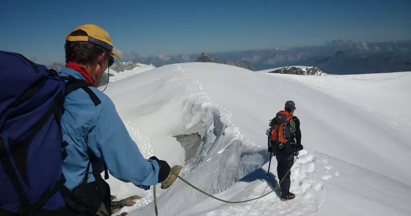 Twee bergbeklimmers met rugzakken en uitrusting, aan elkaar vastgebonden met een touw, trekken over een besneeuwde bergkam onder een strakblauwe hemel.